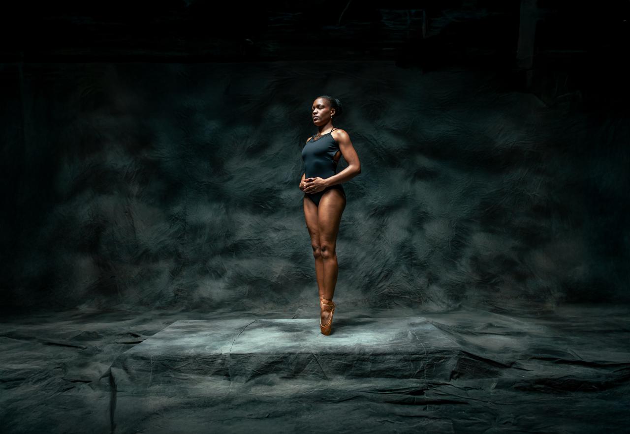 A ballerina stands en pointe on a stone plinth in a dark studio